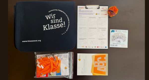 Auf der Abbildung ist das Klassenrat Starterset plus zu sehen. Neben der dunkelblauen Filztasche liegen eine Info-CD zum Klassenrat, Der Mini-Guide in ein Klemmbrett geklemmt, ein Ball aus Gummifäden, ein ABC der Demokratiepädagogik und das Klassenrat Miniset.