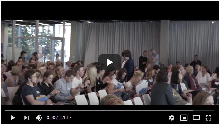 Es handelt sich um den Screenshot eines Videos. In langen Reihen von weißen Stühlen nehmen Personen Platz.