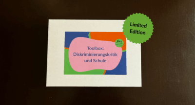 Auf dunklem Hintergrund steht eine flache weiße Box mit einem bunten quadratischen Sticker mit der Aufschrift "Toolbox: Diskriminierungskritik und Schule". Auf dem Bild liegt eine runde Form mit der Aufschrift "Limited Edition".
