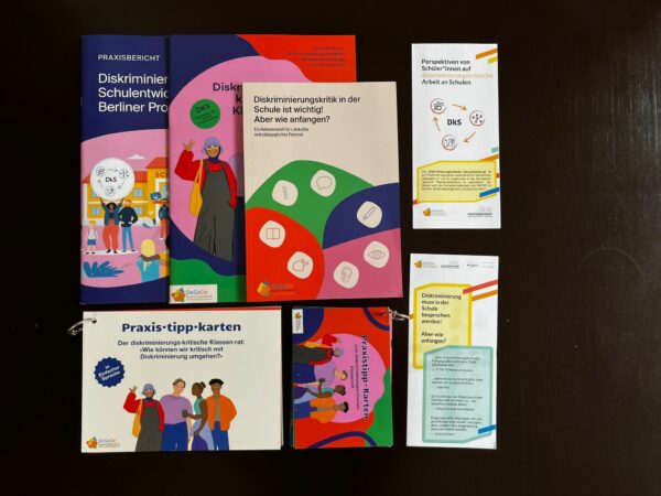 Auf einer dunklen Oberfläche liegt der Inhalt der Toolbox: Der Praxisbericht, der Diskriminierungskritische Klassenrat, das Reflexionsheft, die Praxistippkarten in normaler und einfacher Sprache und die Faltblätter "Schüler*innen-Perspektiven: diskriminierungskritische Projekte an Schulen" und "Zum Erkennen und Vermeiden von Diskriminierung in der Schule".