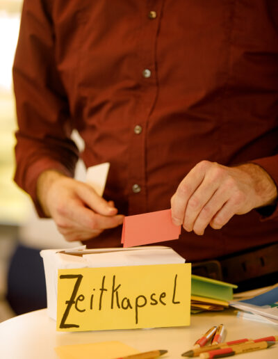 Man sieht eine Person, deren Hand einen Zettel in eine Schachtel mit der Aufschrift 'Zeitkapsel' steckt.